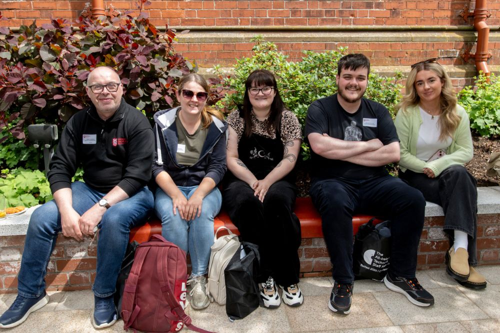 Five conference attendees (two male and three female), sitting in the sunshine on a low brick wall in front of plants at the side of a red brick building